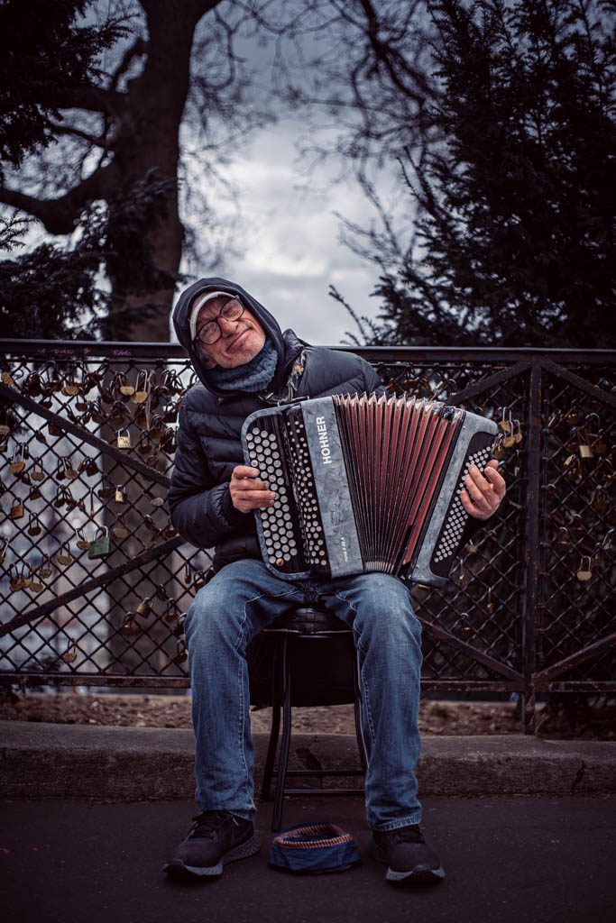 uliczny grajek, starszy mężczyzna grający na harmonii i zbierający pieniądze do kapelusza. W tle płat z zawieszonymi kłódkami 