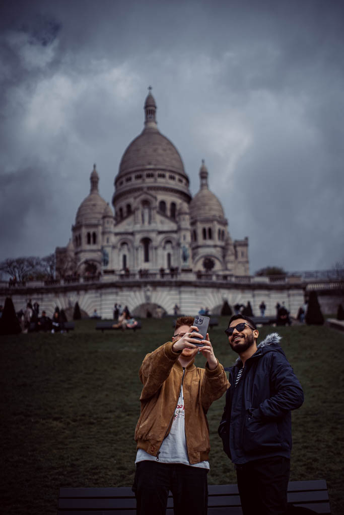 Dwie osoby robiące sobie selfie z Montmartre w tle. Uśmiechy na twarzy i słoneczne okulary pomimo pochmurnej pogody.